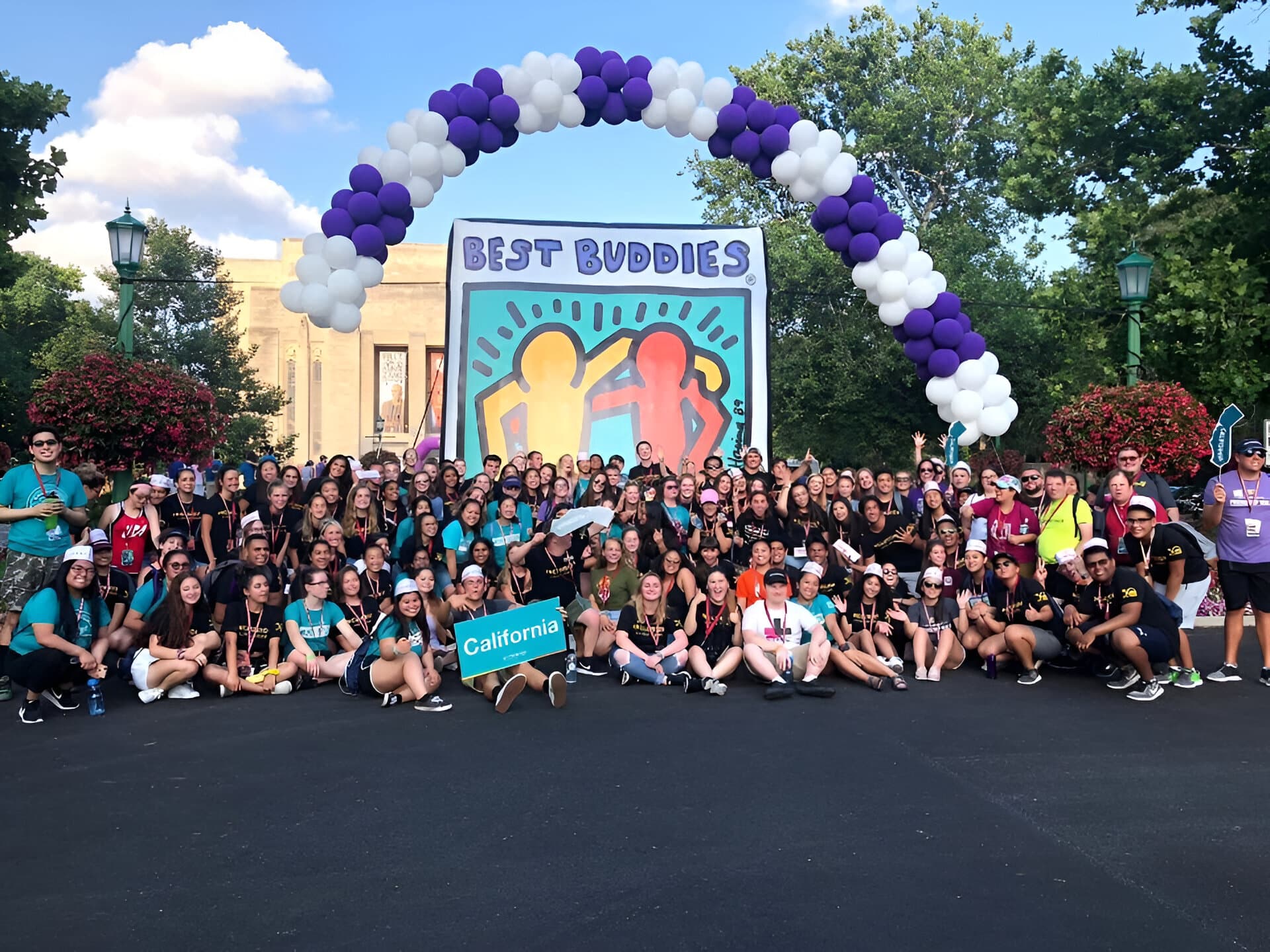 Large group of members posing together in front of a banner and a purple and white ballon arch.