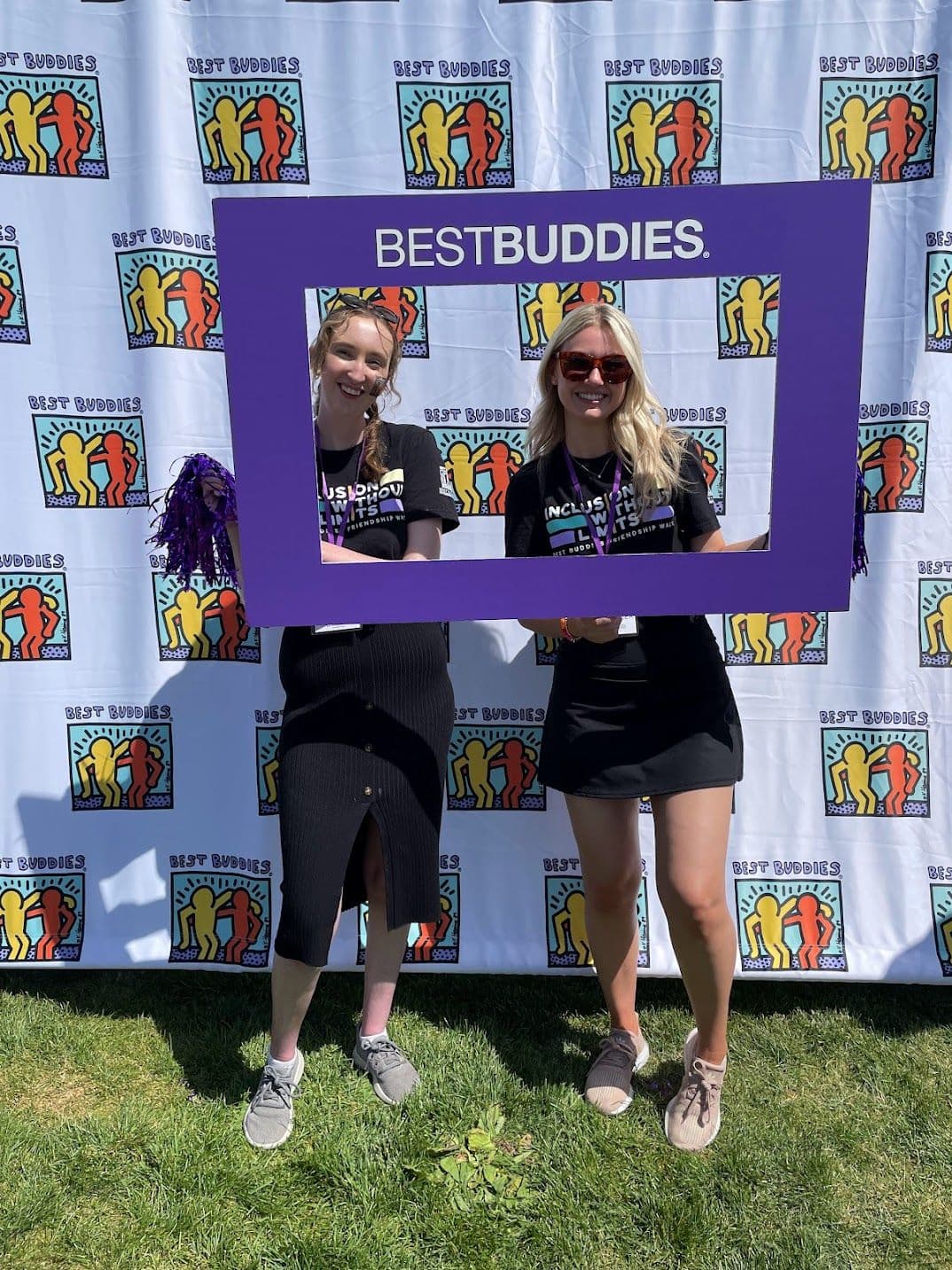 Two women stand in the center of a purple cardboard frame that says "Best Buddies" in white text. They are wearing black Friendship Walk t-shirts.  In the background is a white curtain with the Best Buddies logo in a repeating pattern.