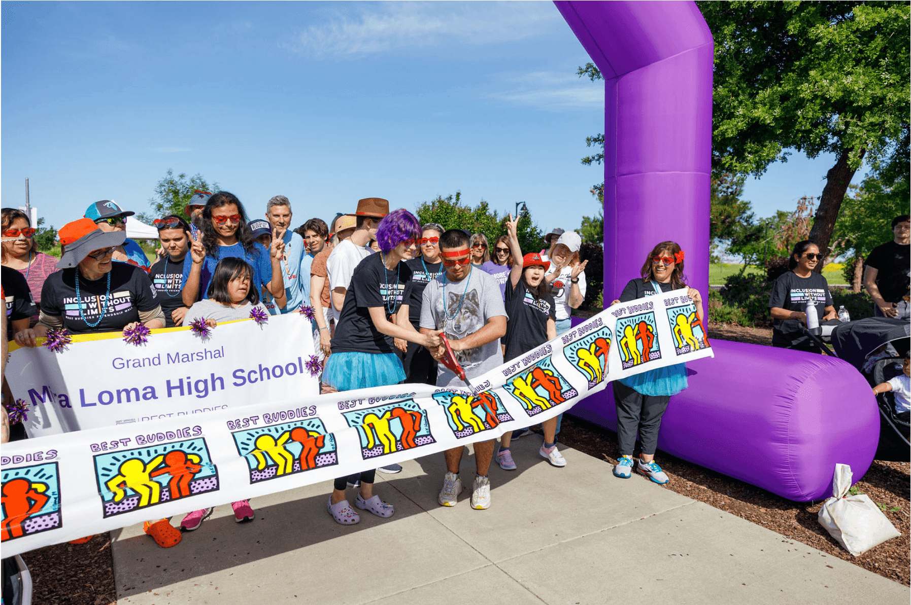 A crowd of people is gathered behind an inflatable purple archway. Two people in the center of the photo are cutting a ribbon patterned with the Best Buddies logo. They are beginning the walk course. 