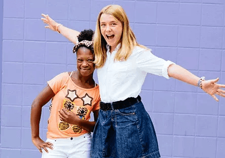 A buddy pair, both girls, stand in front of a purple wall. They are smiling and posing together.