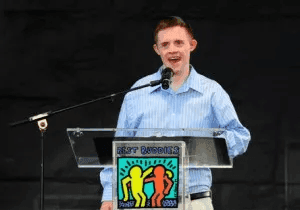 A Best Buddies Ambassador stands at a podium and speaks into a microphone. He is wearing a blue button-down shirt and tan slacks.