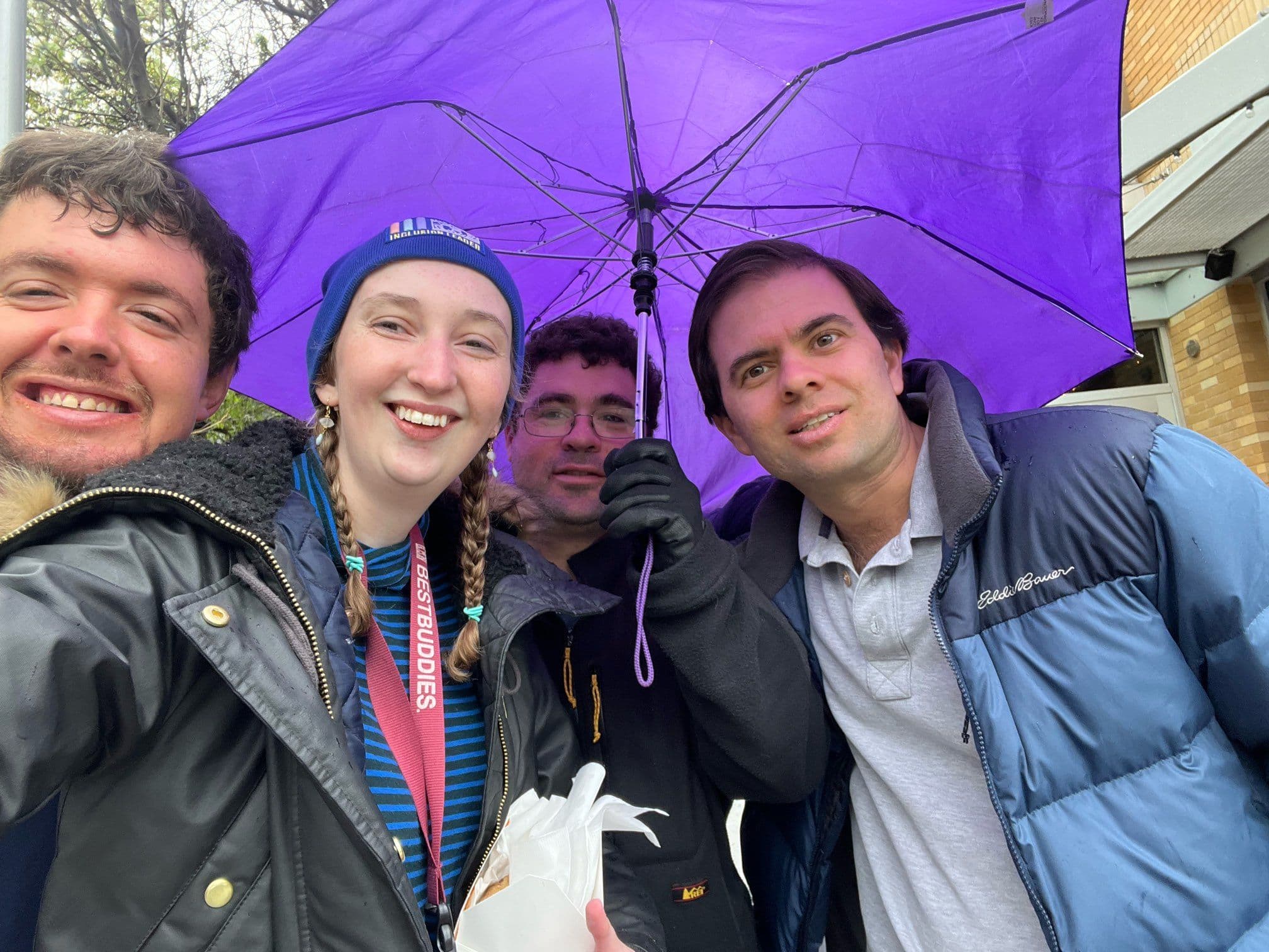 Four Best Buddies members are huddled under an umbrella and smiling at the camera.