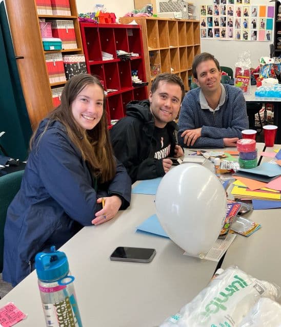 Five Best Buddies members are sitting around a table and smiling at the camera. Multicolored construction paper, scissors, cotton balls, and other craft supplies are on the table.