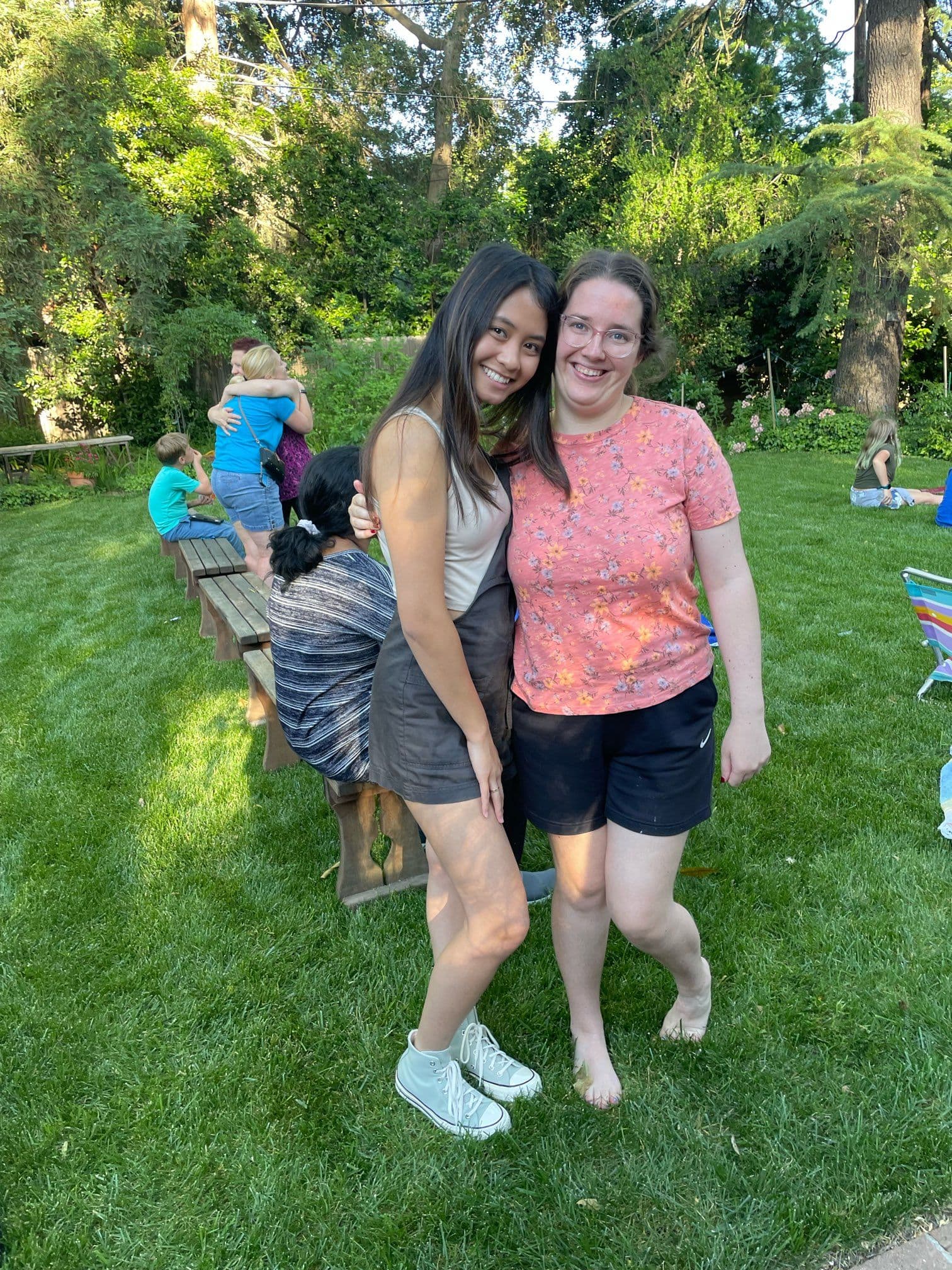 Two Best Buddies members, both girls, smile at the camera and pose together. In the background is a lush green lawn and trees.